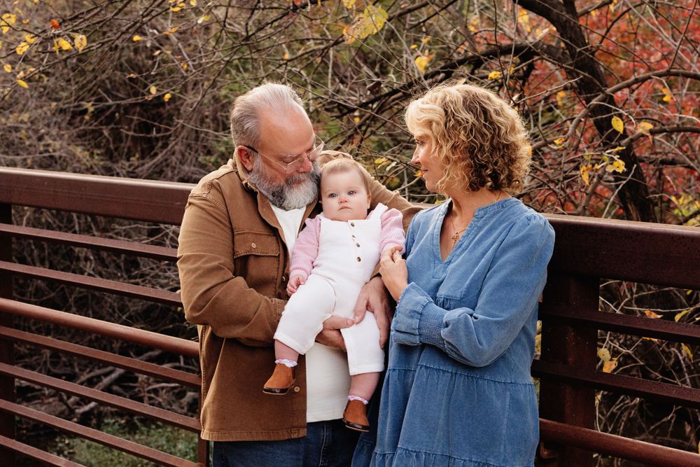 A couple shares a tender moment with their baby on a rustic bridge during autumn with colorful foliage in the background.