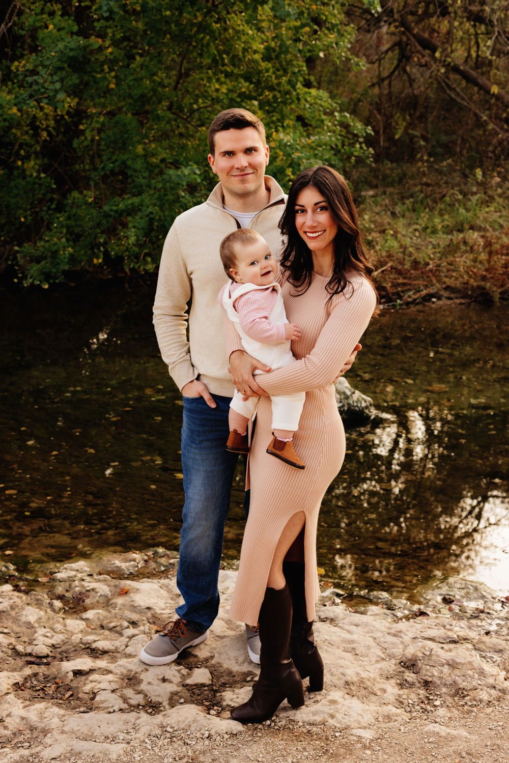 A young family poses together outdoors in autumn by a calm stream wearing coordinated neutral colored outfits.