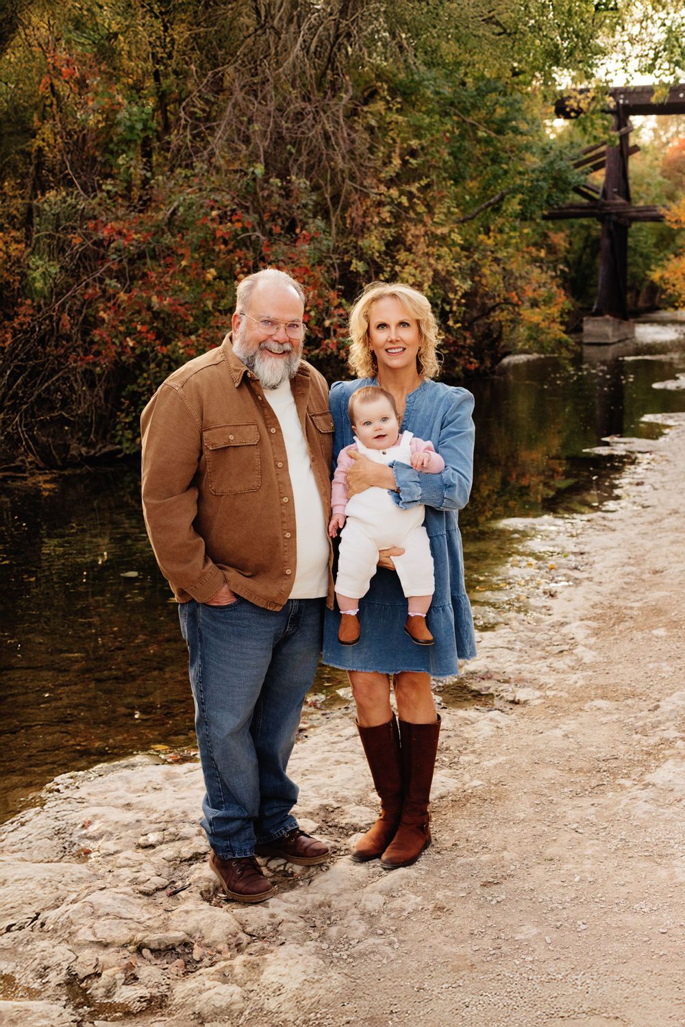 Three generations pose on an autumn path by a tranquil stream surrounded by fall foliage and trees.