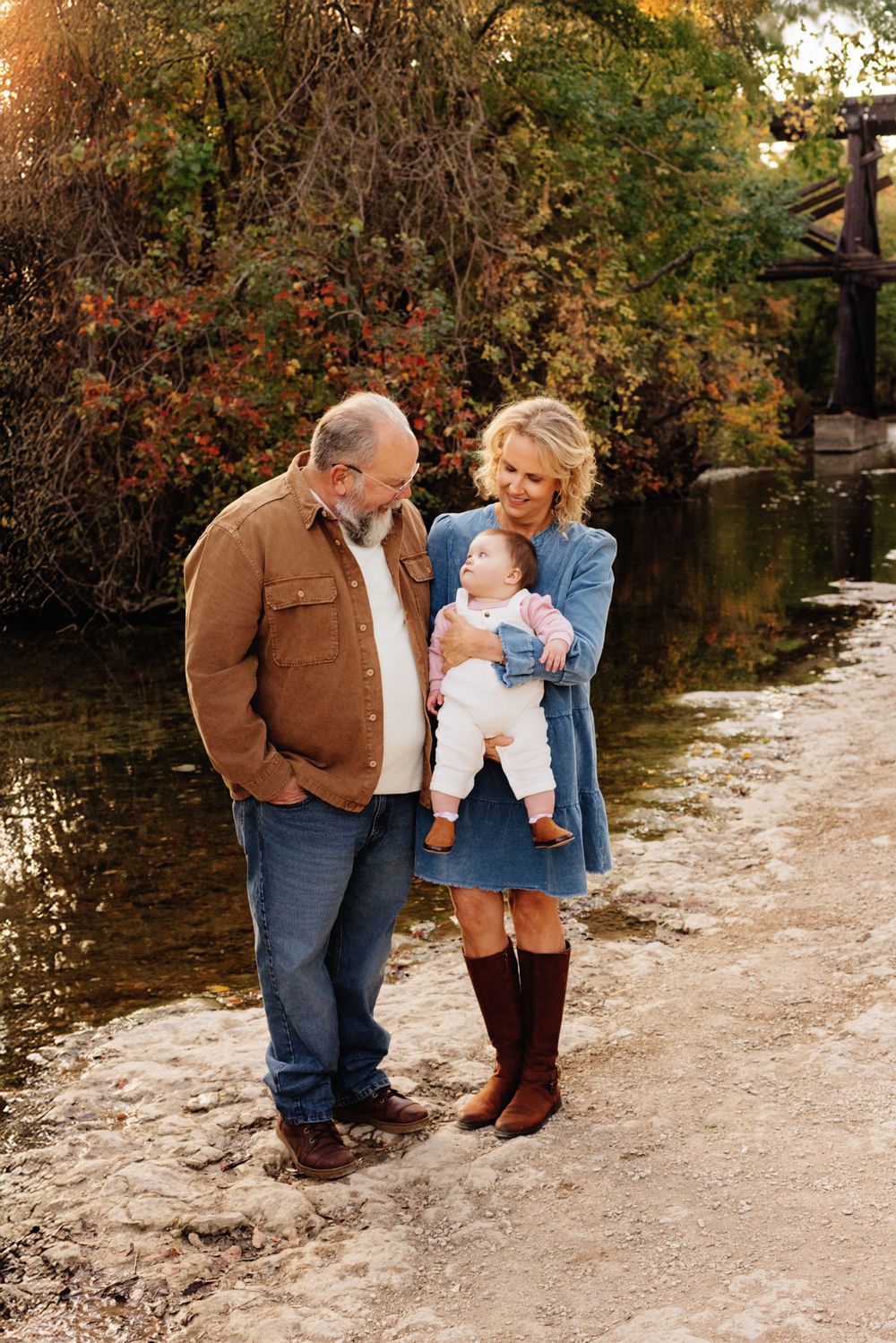 A family stands together on a dirt path by a pond surrounded by autumn foliage on a sunny fall day.