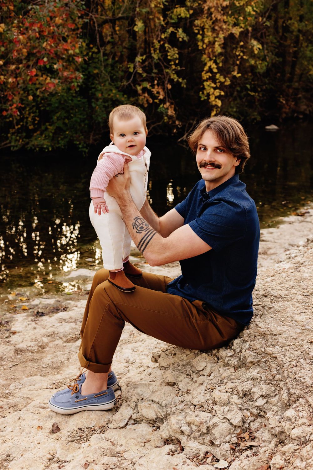 An adult in a navy shirt and brown pants sits with a young child in pink by a waterside with autumn foliage in background.