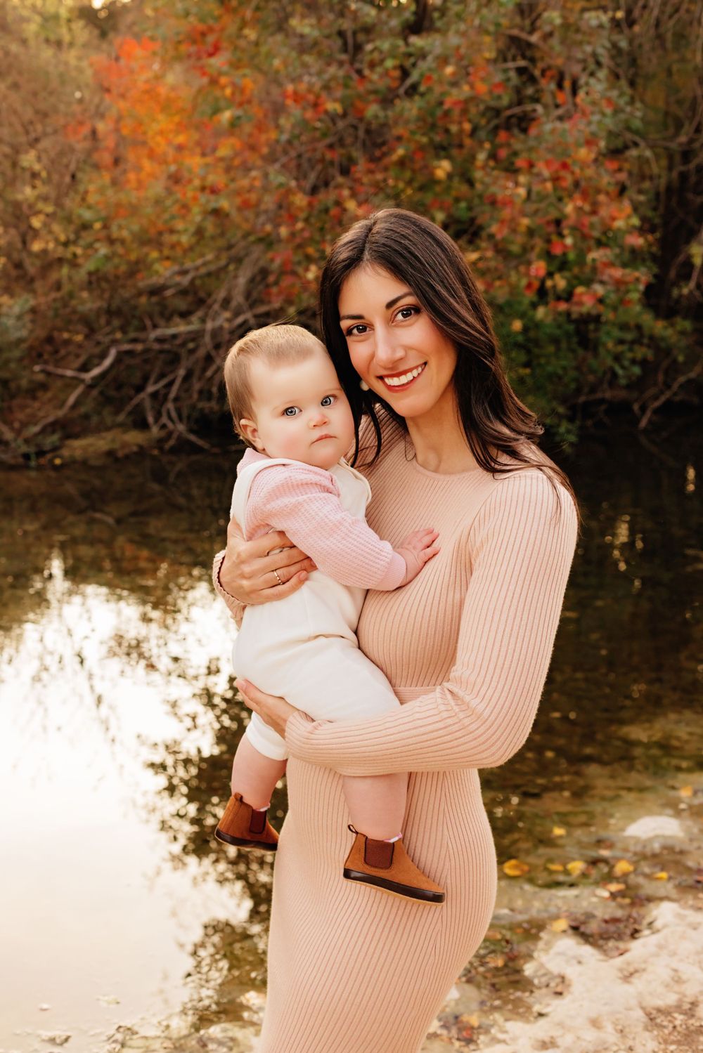 A heartwarming fall portrait of mother and baby in matching pink outfits standing by a pond with autumn foliage.