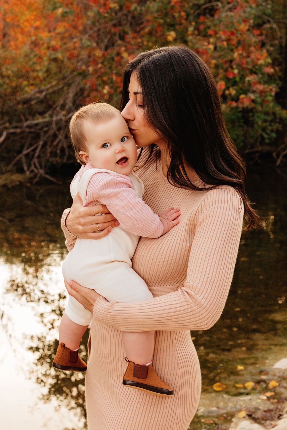 An intimate moment between mother and baby captured against vibrant fall colors reflecting in a peaceful pond.