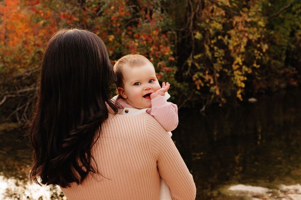 Someone in a peach sweater holds a baby while standing near a pond with colorful autumn foliage in the background.