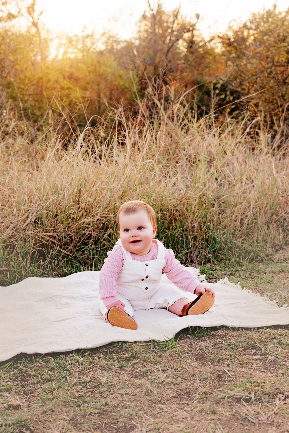 Sweet little one wearing a pink cardigan and white overalls sits on blanket in golden fall field at sunset.