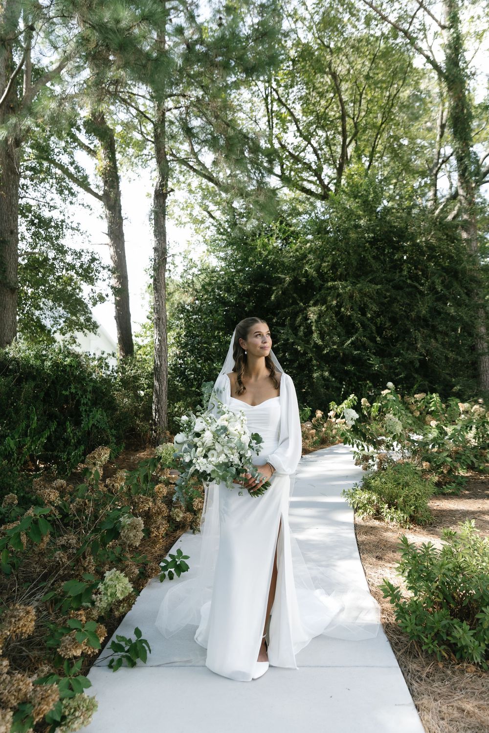 Elegant bridal portrait in white dress with bouquet against natural garden backdrop.