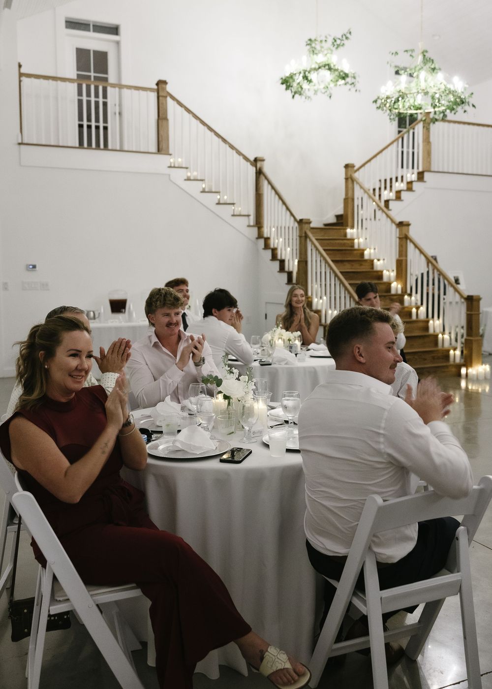 Wedding reception guests seated at elegant round tables with white linens in a bright, airy venue with wooden staircase.