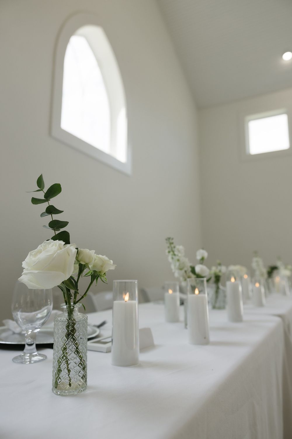 White roses in glass vases and candles decorate elegant tables at a wedding reception.
