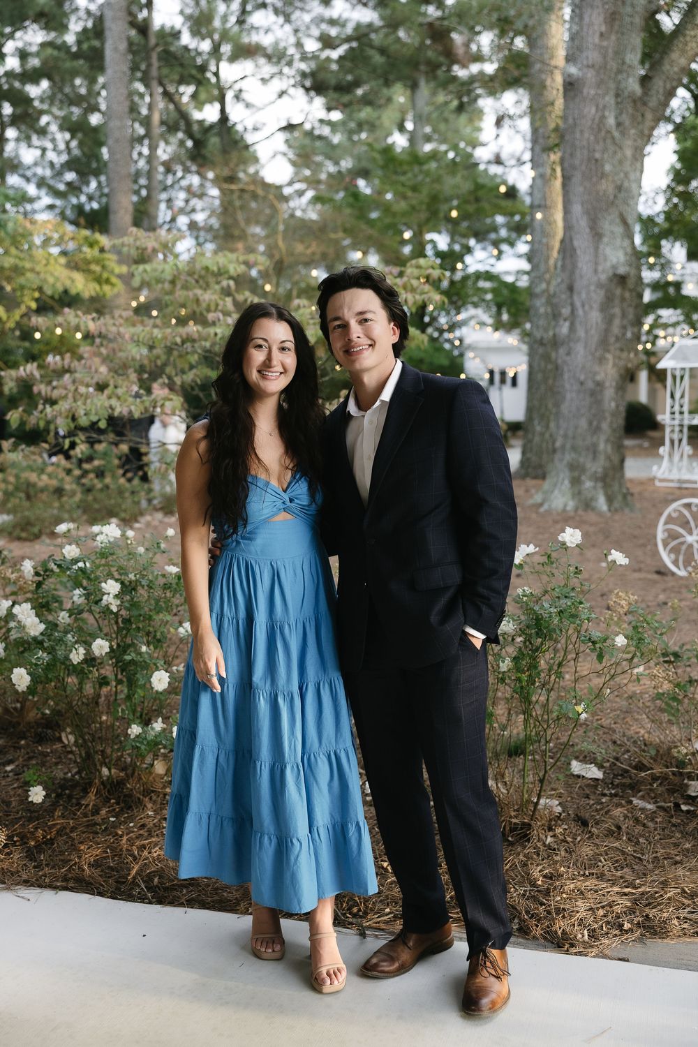 A couple poses together outdoors wearing blue dress and black suit among trees and flowers.