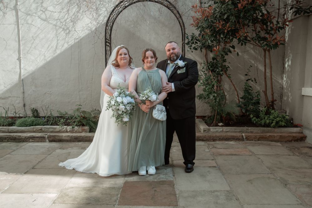 Wedding party in formal attire stands together against a stone wall with bouquets and greenery.