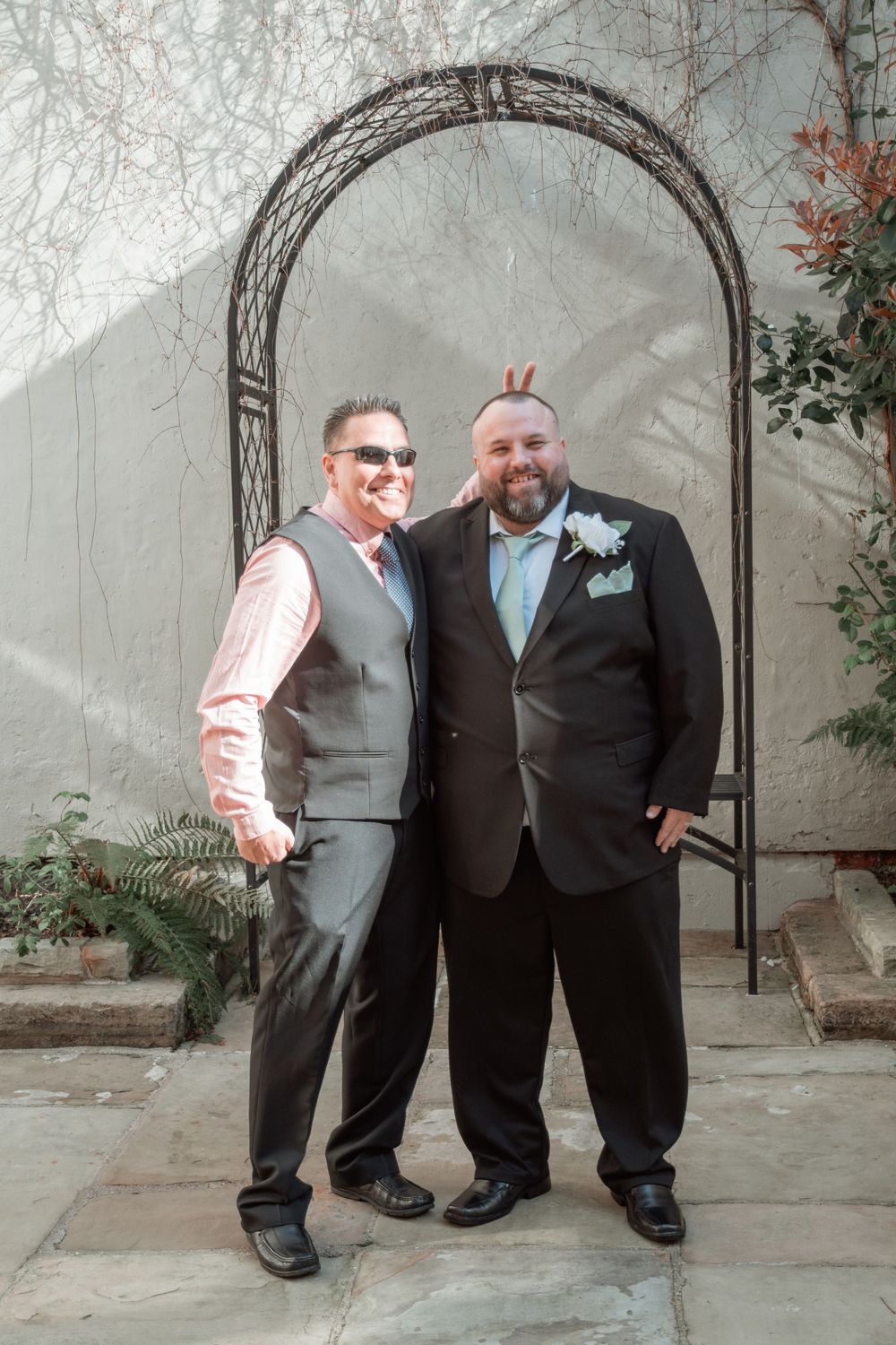 Two wedding attendees in formal attire pose together under a decorative metal garden arch against a light wall.