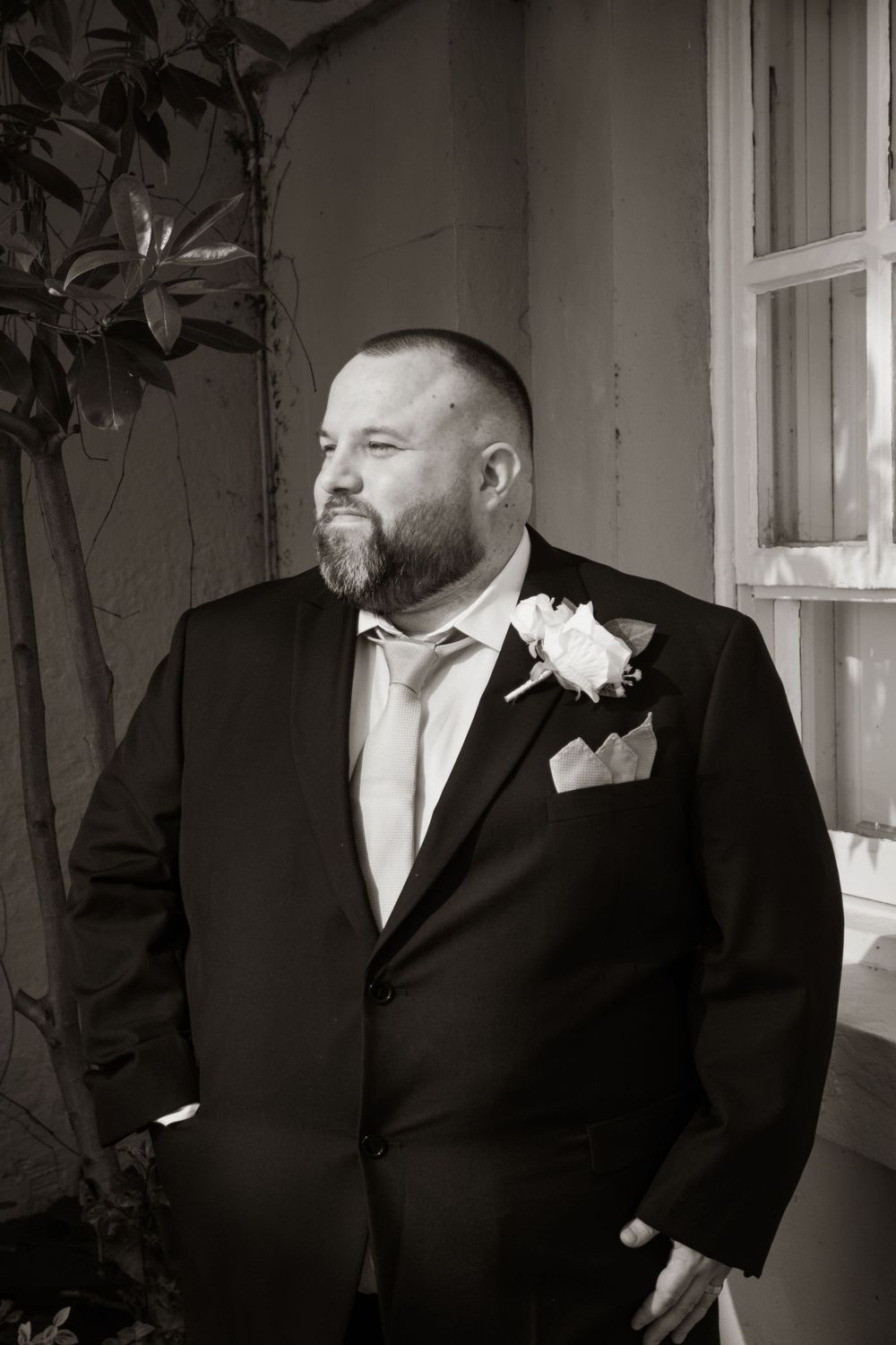 Black and white portrait of groom in formal tuxedo with boutonniere and beard looking to side.