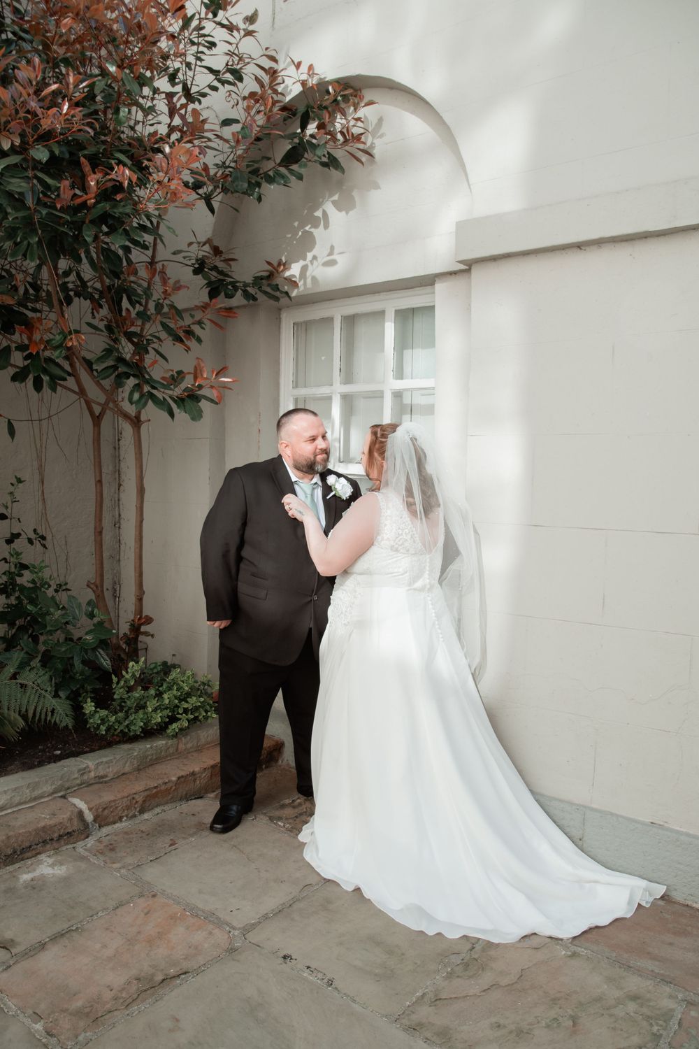 A couple in formal wedding attire stand together in an outdoor alcove with greenery and white walls.