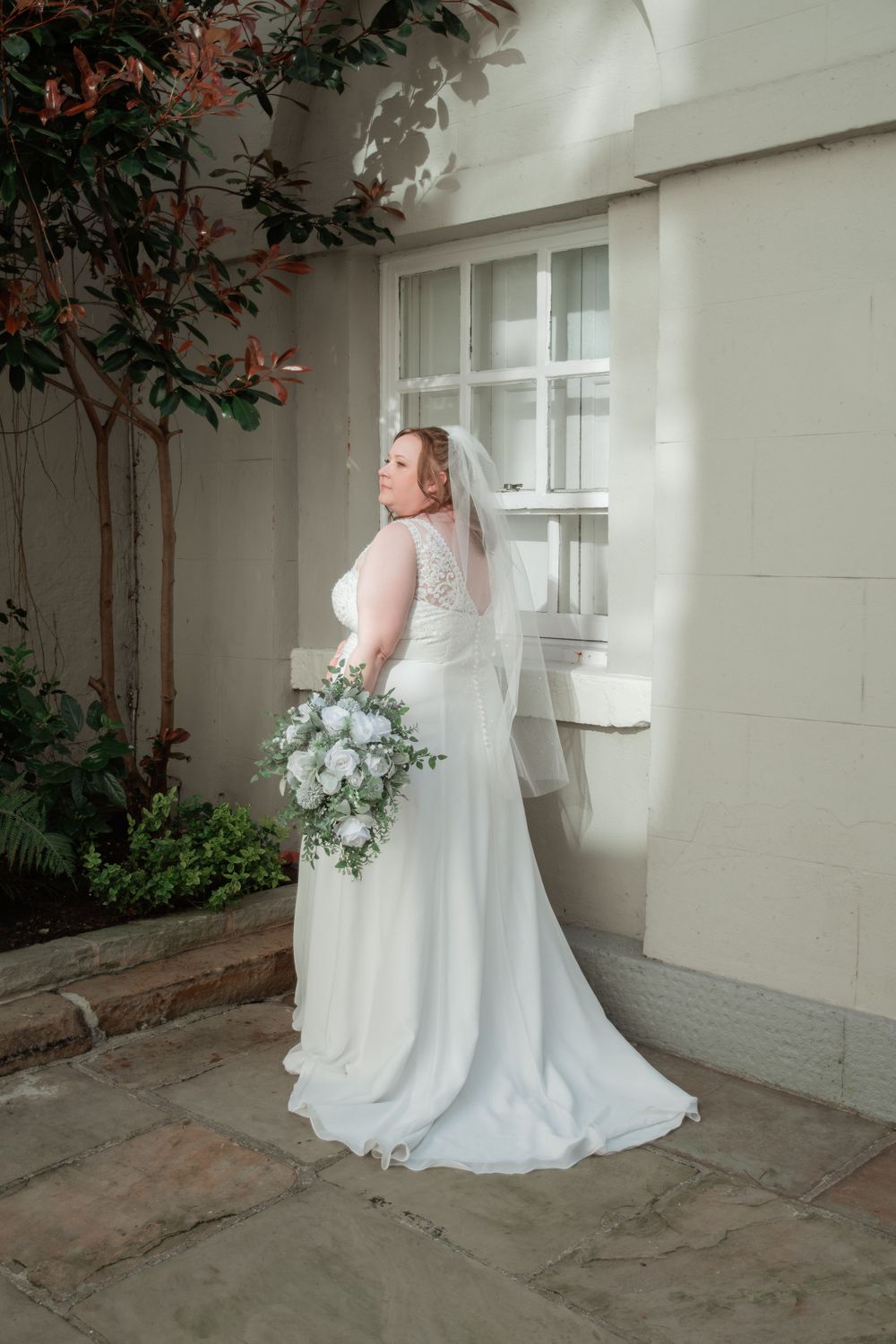 A bride in a flowing white gown and veil poses with a white floral bouquet against a cream colored wall outdoors.