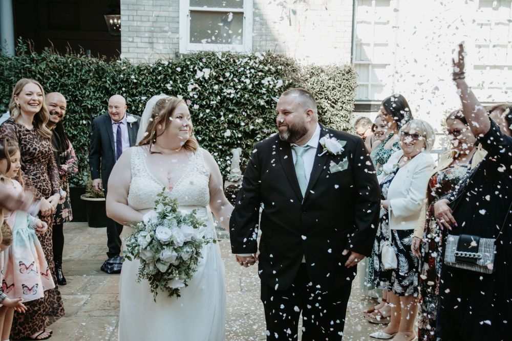 Wedding guests toss white confetti at a couple during an outdoor celebration at a venue with greenery.