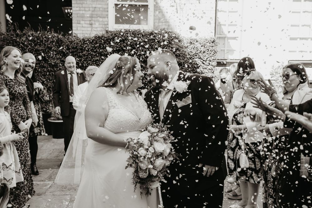 Black and white photo of newlyweds celebrating amid a shower of confetti with wedding guests.