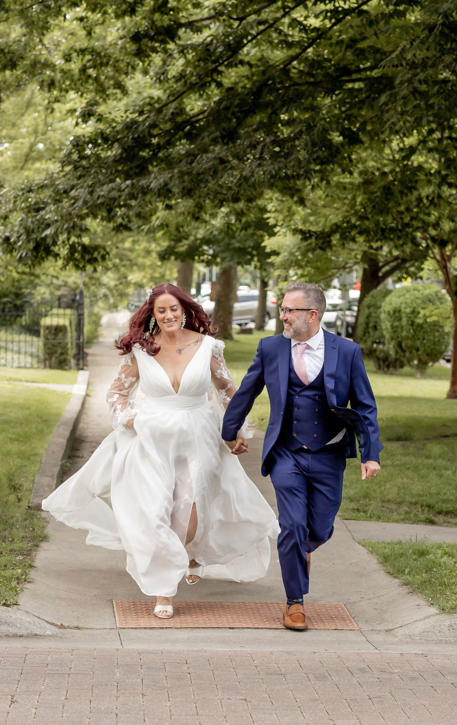 Joyful newlywed couple running hand-in-hand on a tree-lined sidewalk, bride in white lace gown and groom in navy suit.