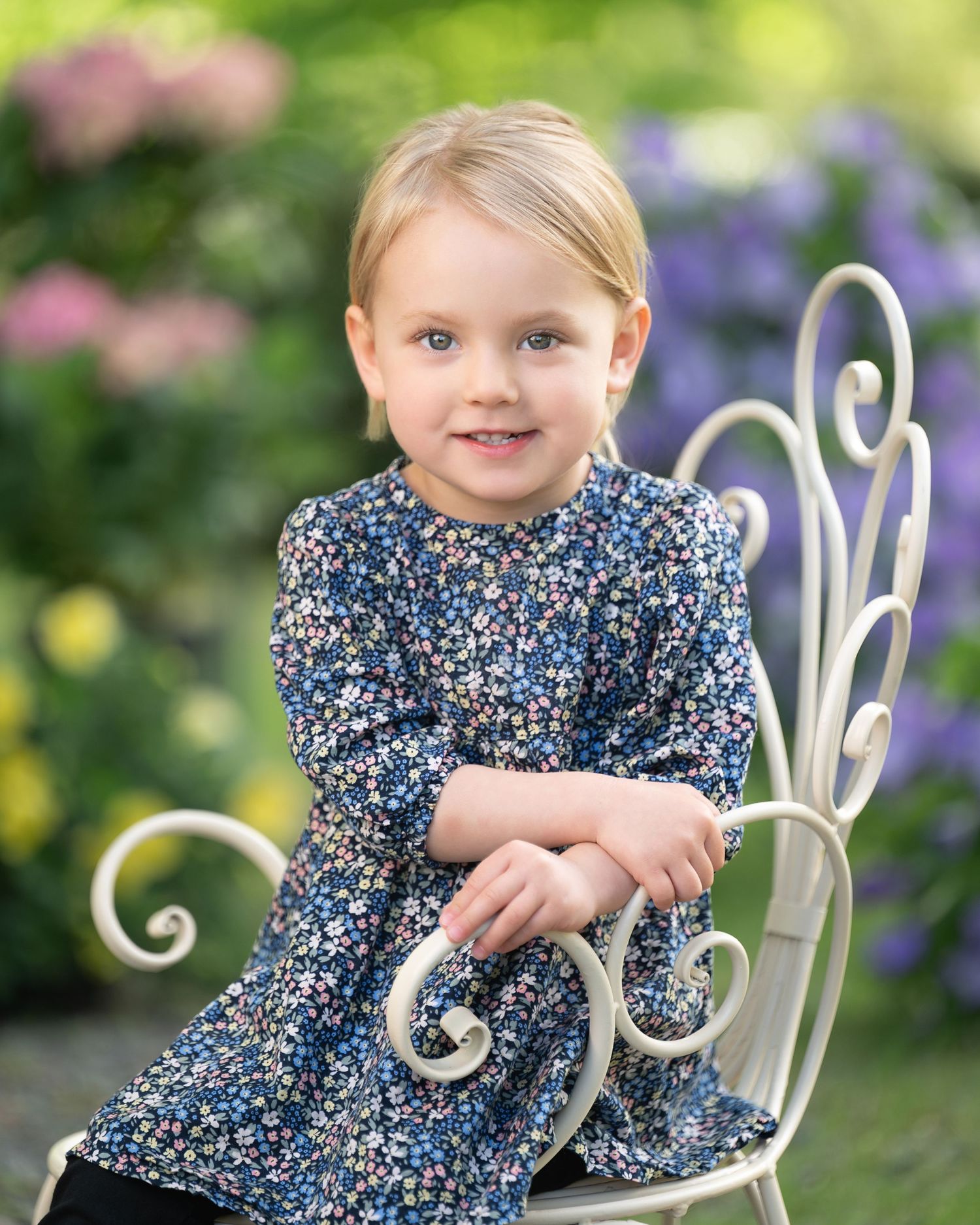 Child in a floral dress with pink bow poses on white garden chair surrounded by colorful flowers in outdoor setting.