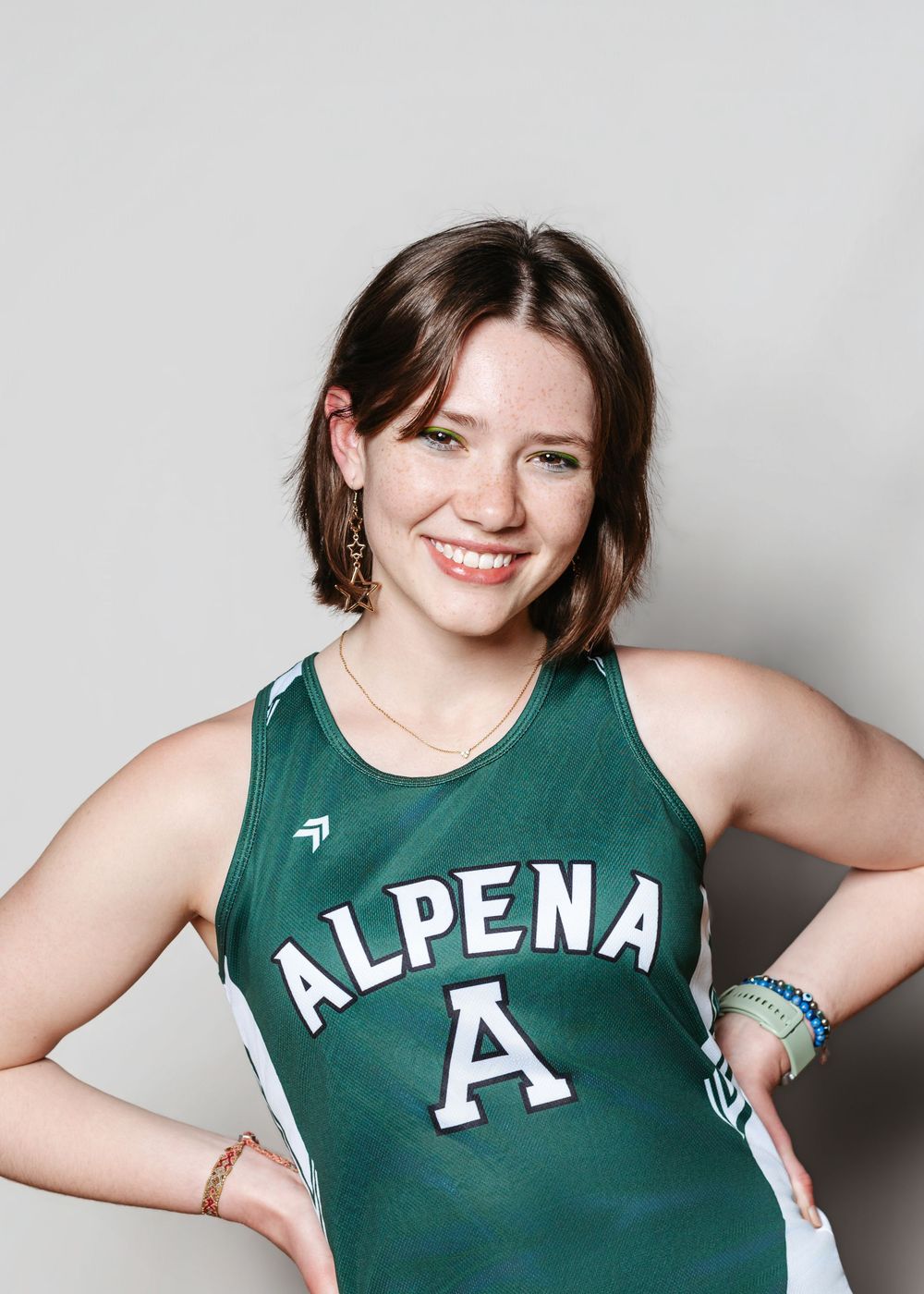 Young woman smiling in green Alpena basketball jersey, hands on hips, against light gray background.
