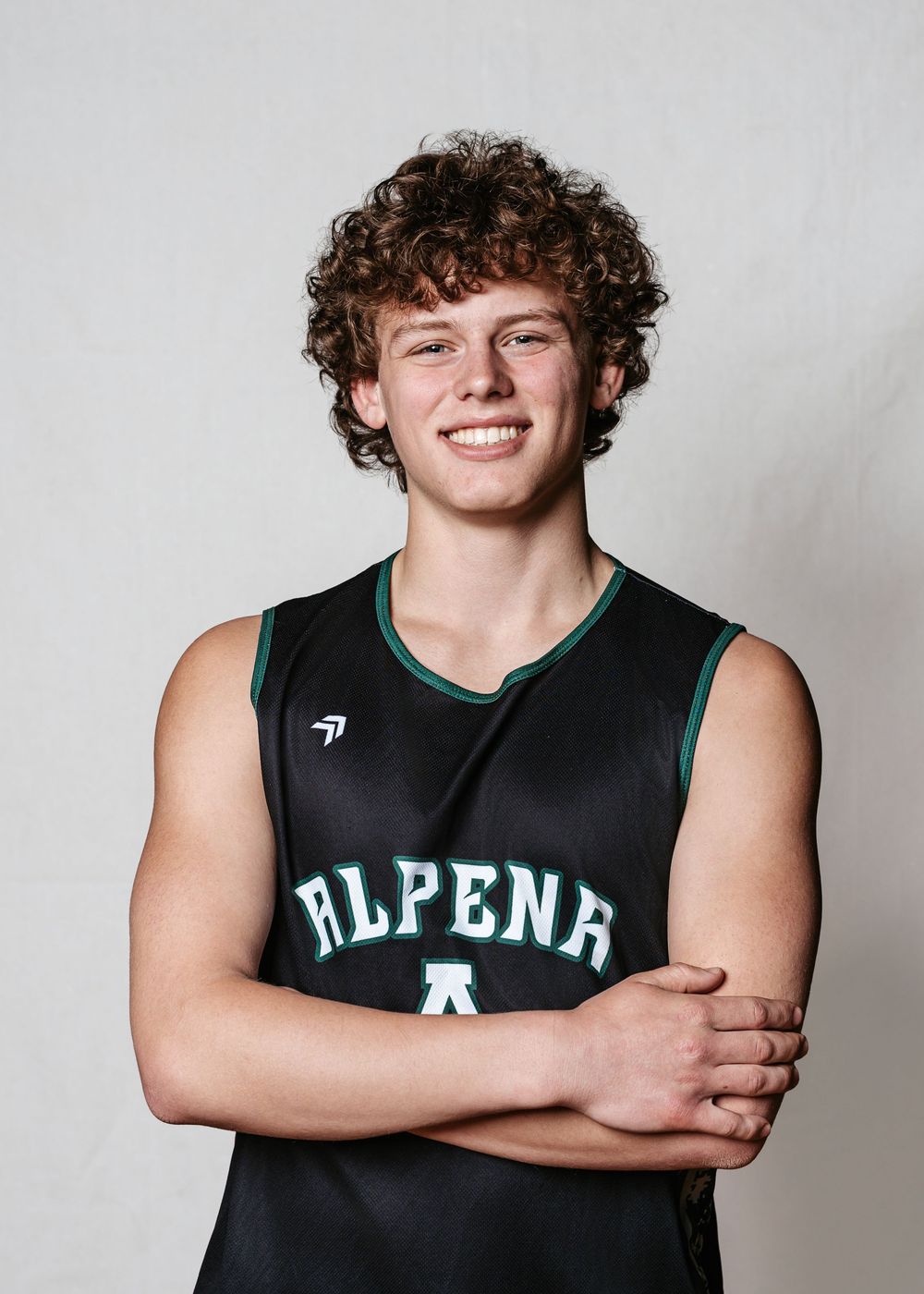 Young male athlete in Alpena black basketball jersey smiling with arms crossed for team photo.