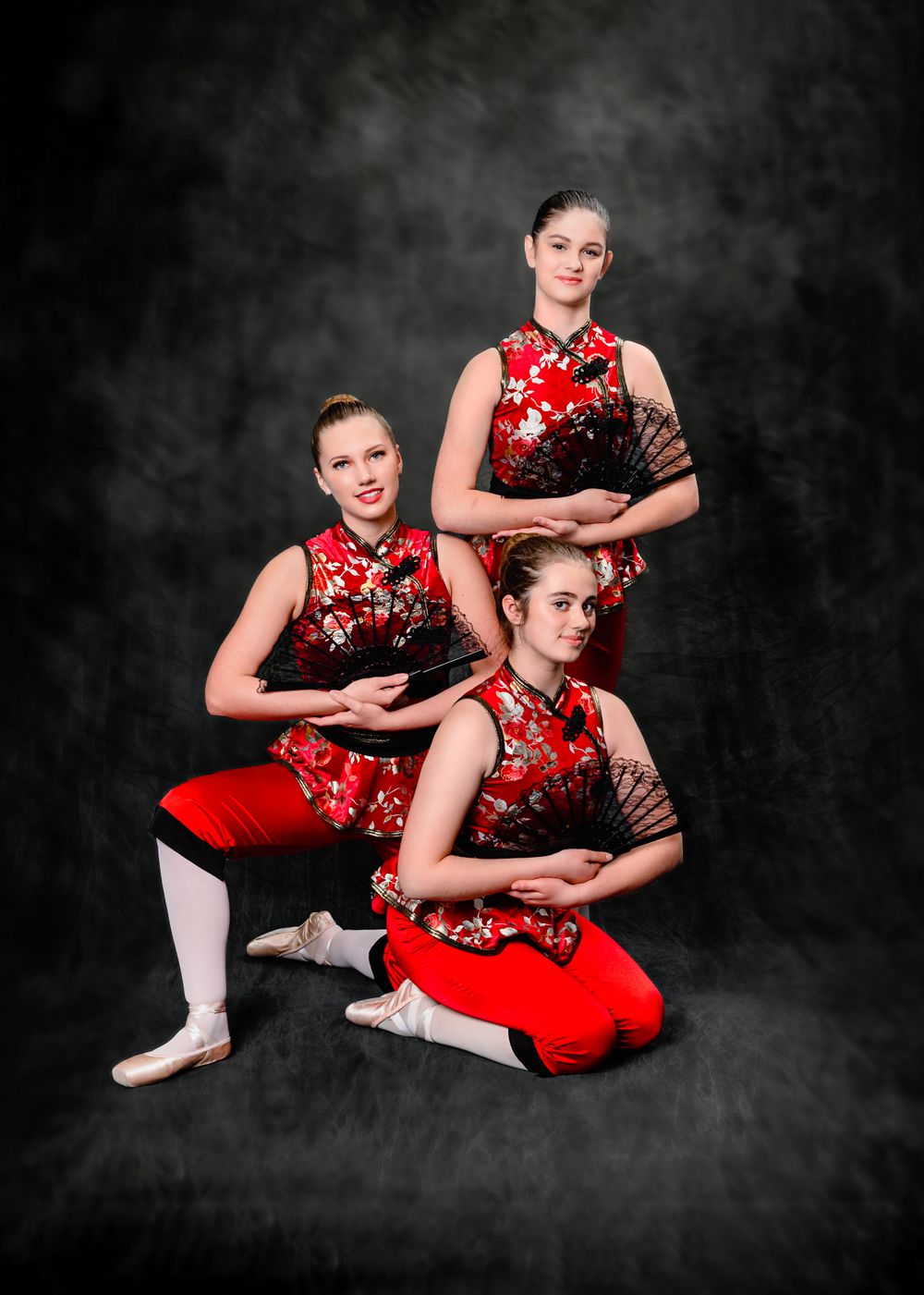 Three dancers in red and black mandarin collar costumes pose elegantly with fans against a dark studio backdrop.