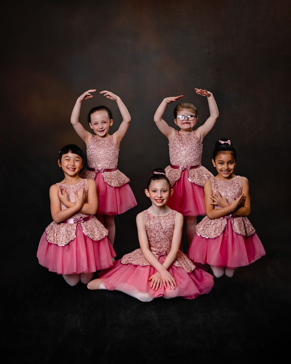 Five young dancers in matching pink dresses and costumes pose together for a dance recital portrait against dark backdrop.
