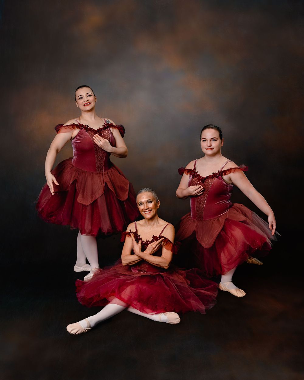 Three ballet dancers in burgundy romantic tutus pose gracefully against a dark dramatic backdrop.