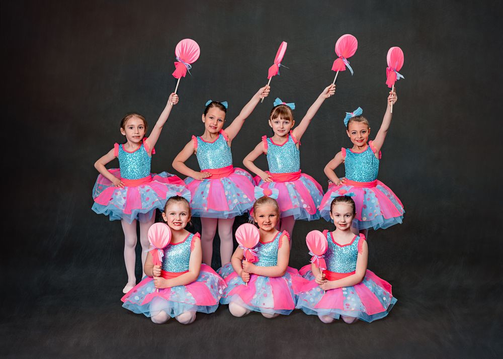 Young dancers in turquoise leotards and pink tutus pose with matching pink props during a dance recital performance.
