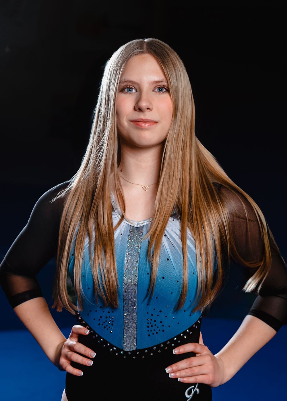Athlete poses in sparkly blue performance leotard with black mesh sleeves against dark backdrop.