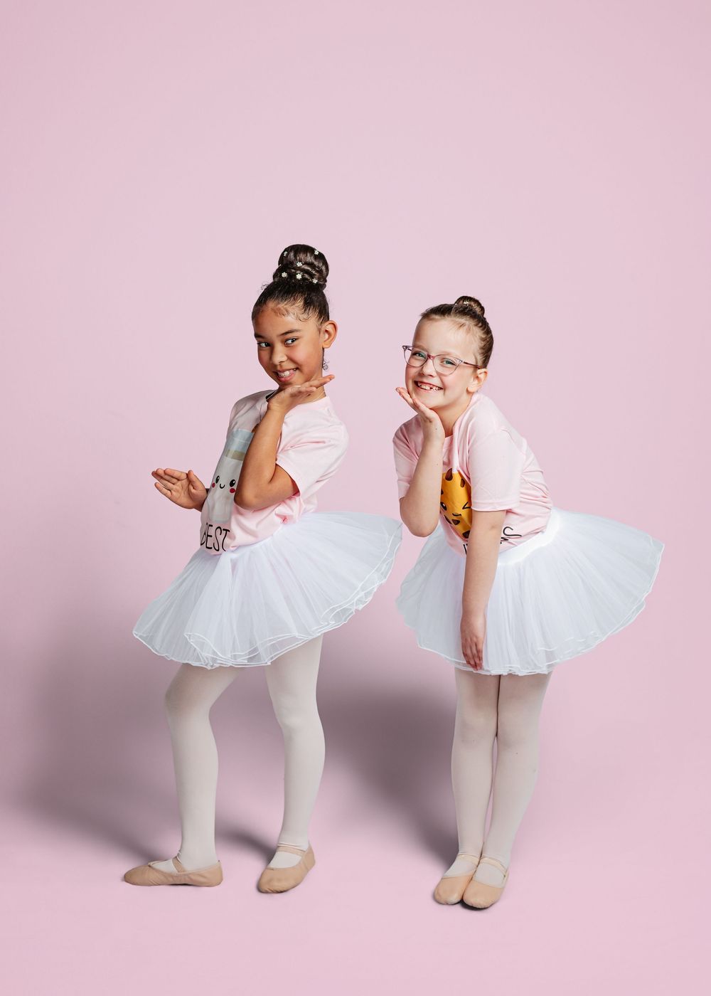 Two young ballet dancers in white tutus and tights share a funny moment against a pink backdrop.