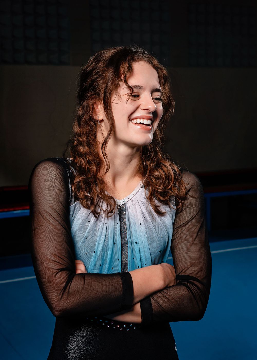 Athlete in blue and black performance outfit with mesh sleeves smiles brightly while standing in a gymnastics facility.