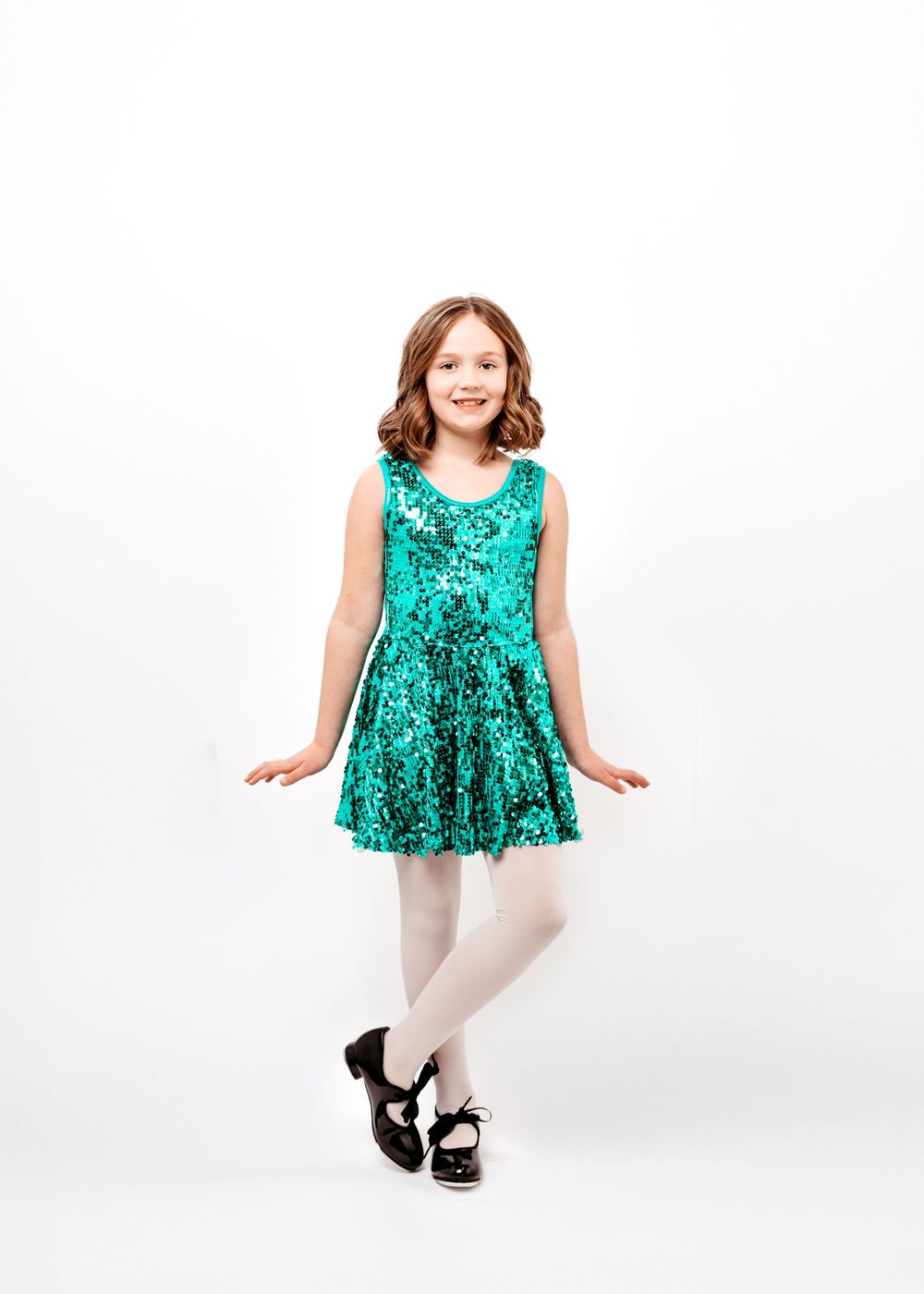 Child dancer in sparkly emerald green dress and white tights strikes a graceful pose against white background.