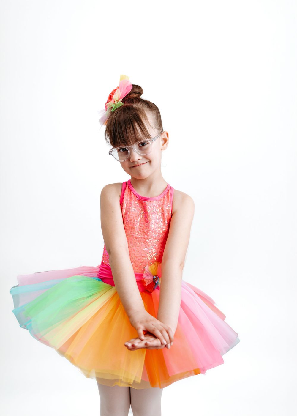 Young dancer in sparkly pink leotard and colorful rainbow tutu performs graceful pose against white backdrop.