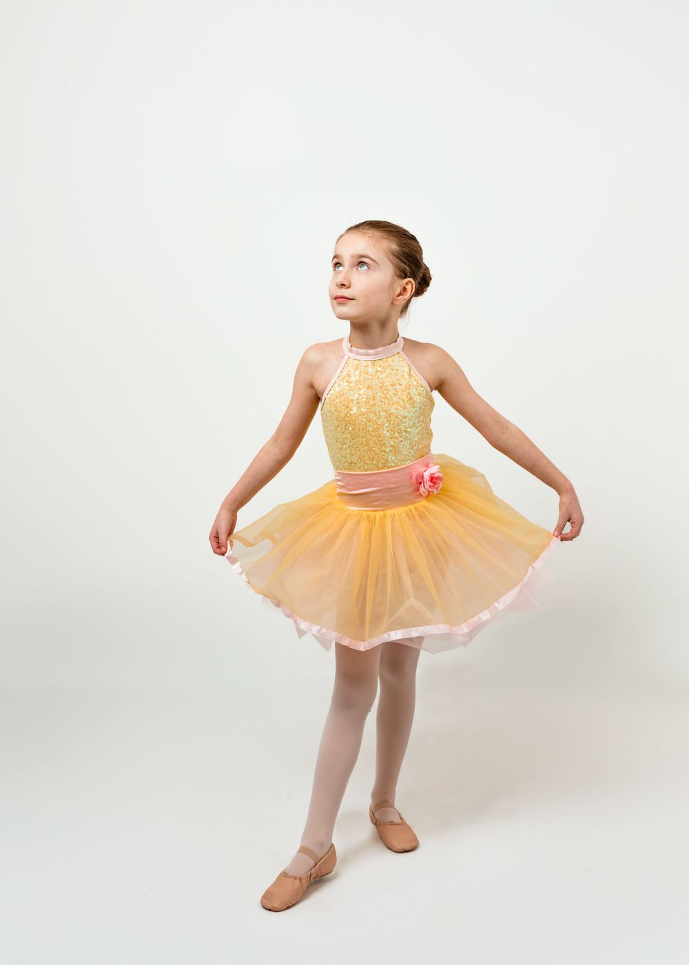 Young dancer in yellow sequined leotard and peach tulle tutu stands gracefully in first position against white backdrop.