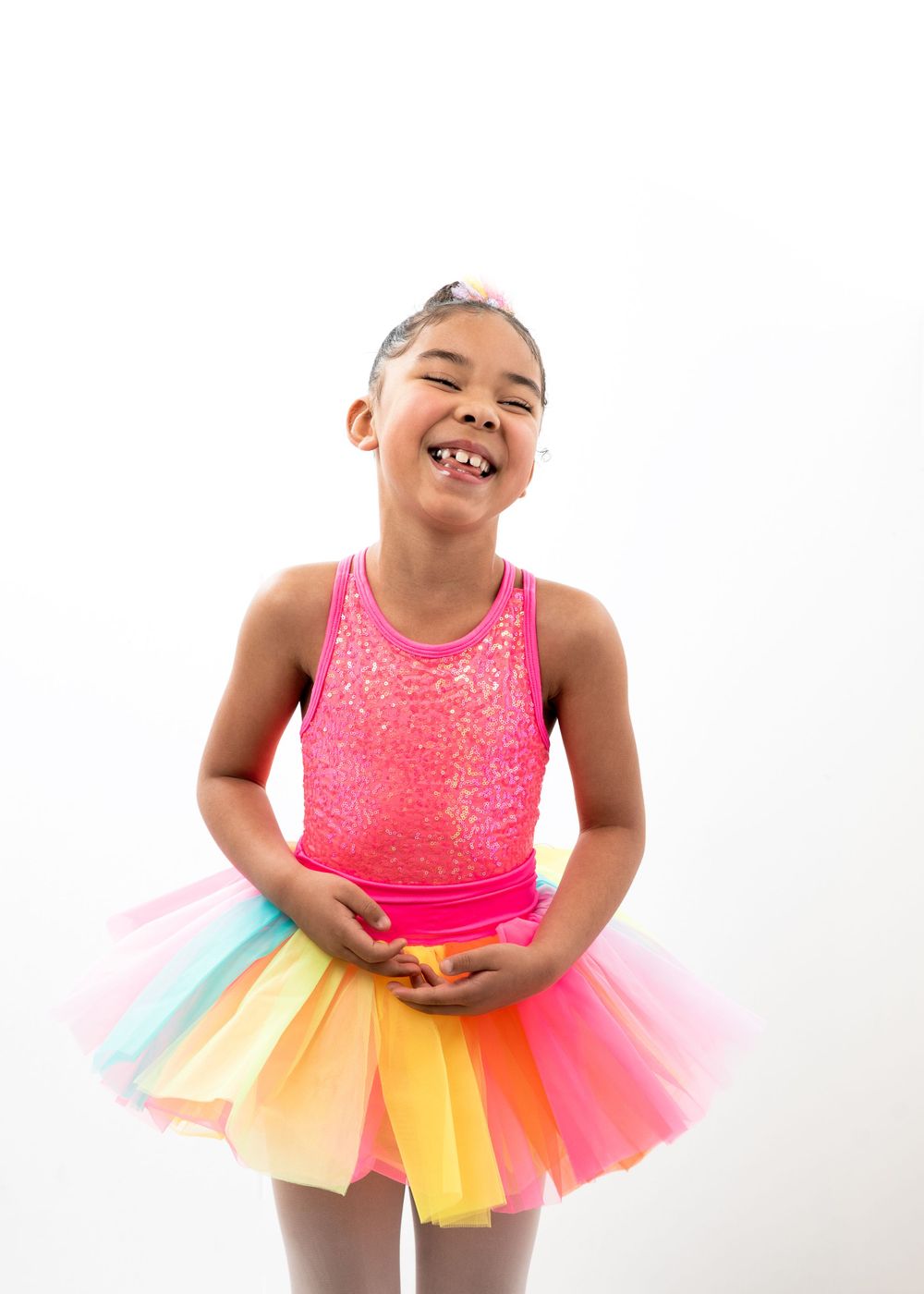 A young dancer wearing a bright pink leotard and rainbow tutu performs with a joyful expression against a white backdrop.