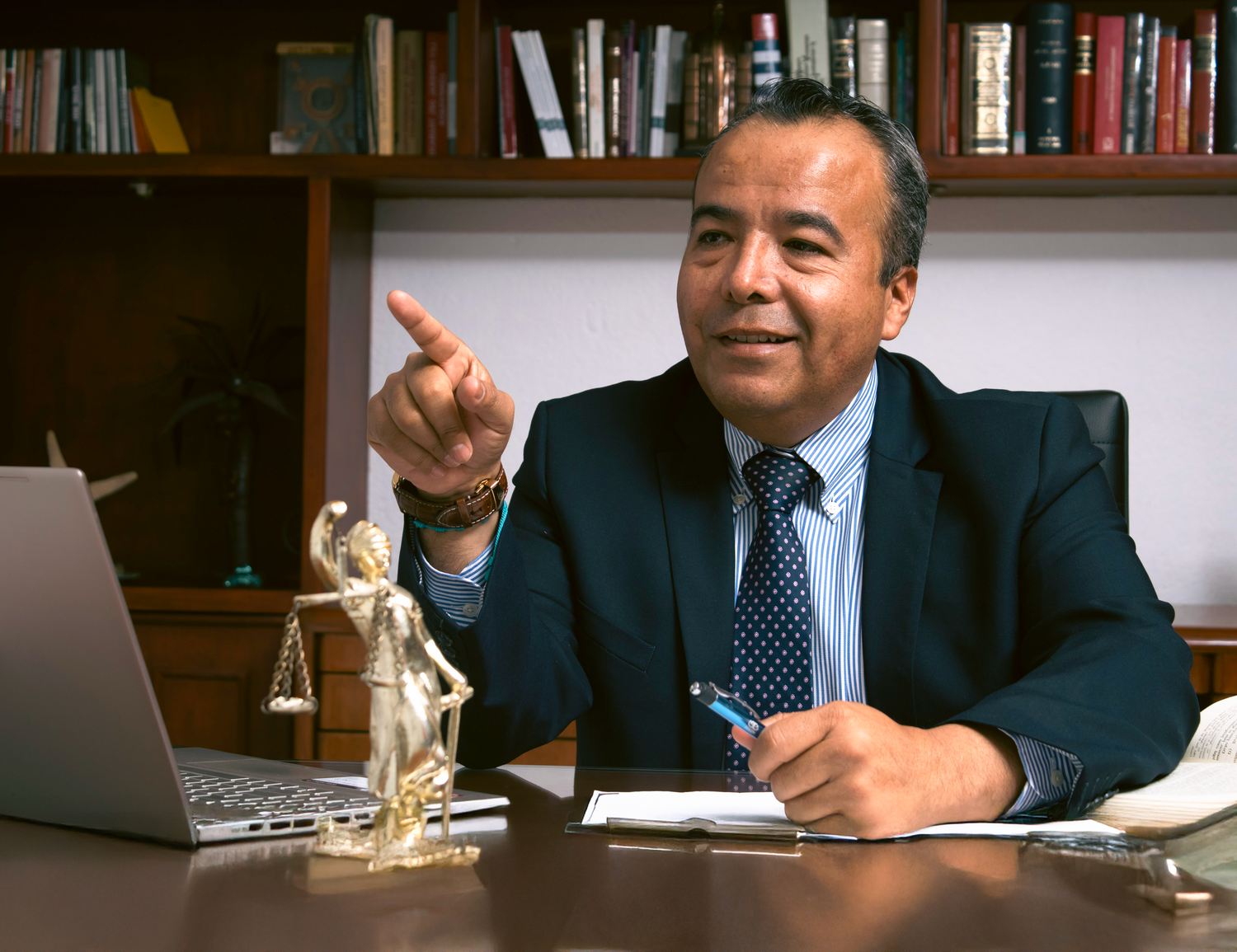 A confident businessman in a suit gestures while seated at a desk with a laptop and Lady Justice statue.