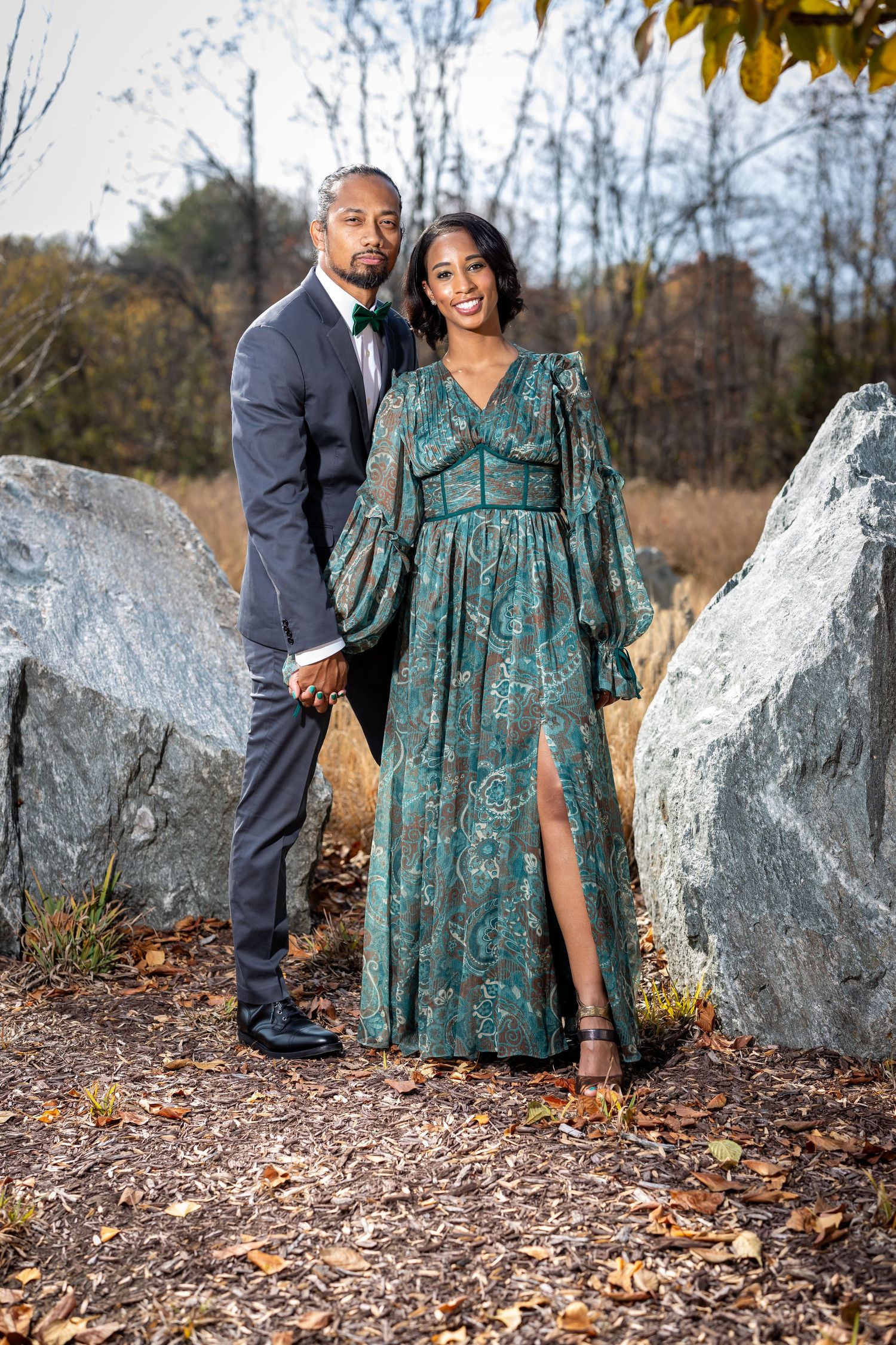 Elegant couple in formal attire posing between large boulders in a natural outdoor setting.