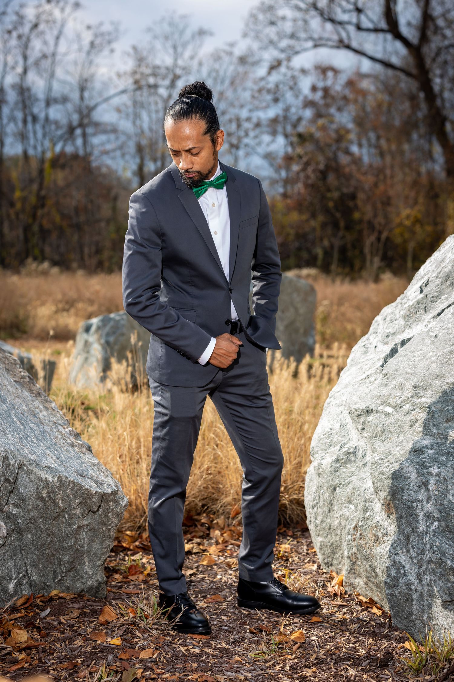 Formal portrait of a suited figure with green bowtie standing between large grey boulders.