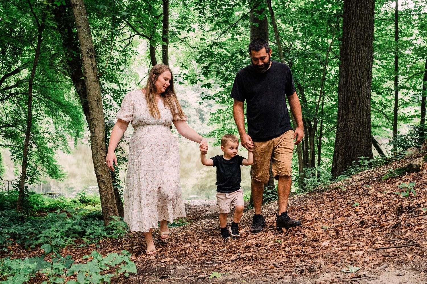 A couple walks hand in hand with their small child along a wooded forest trail covered in pine needles and leaves.
