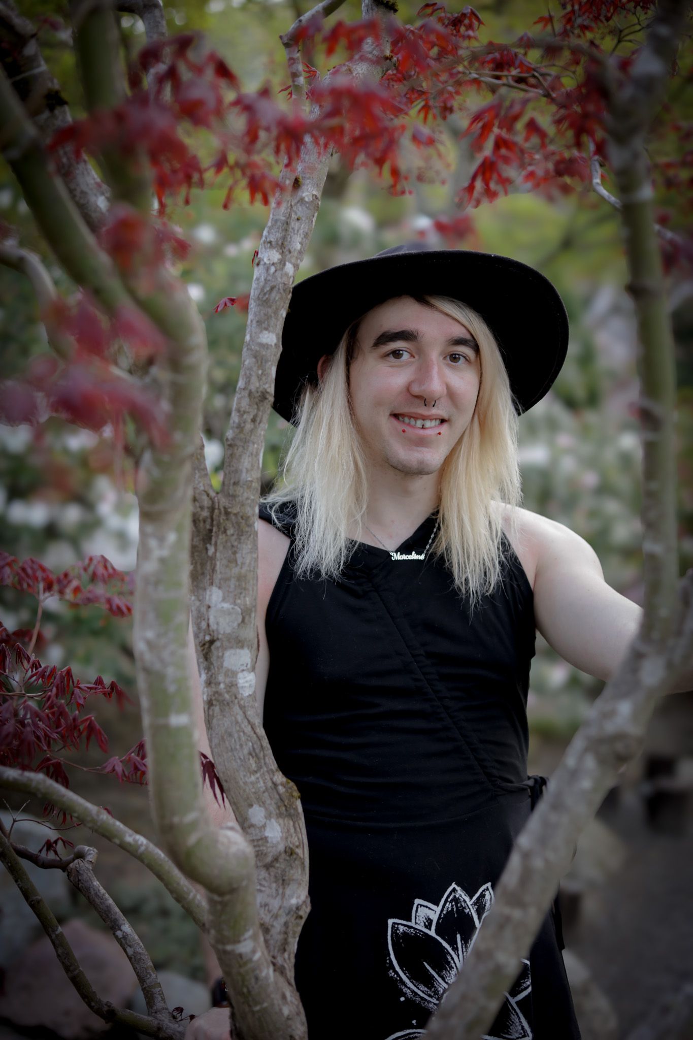 A person wearing a black hat and tank top poses near autumn foliage and pink blossoms.