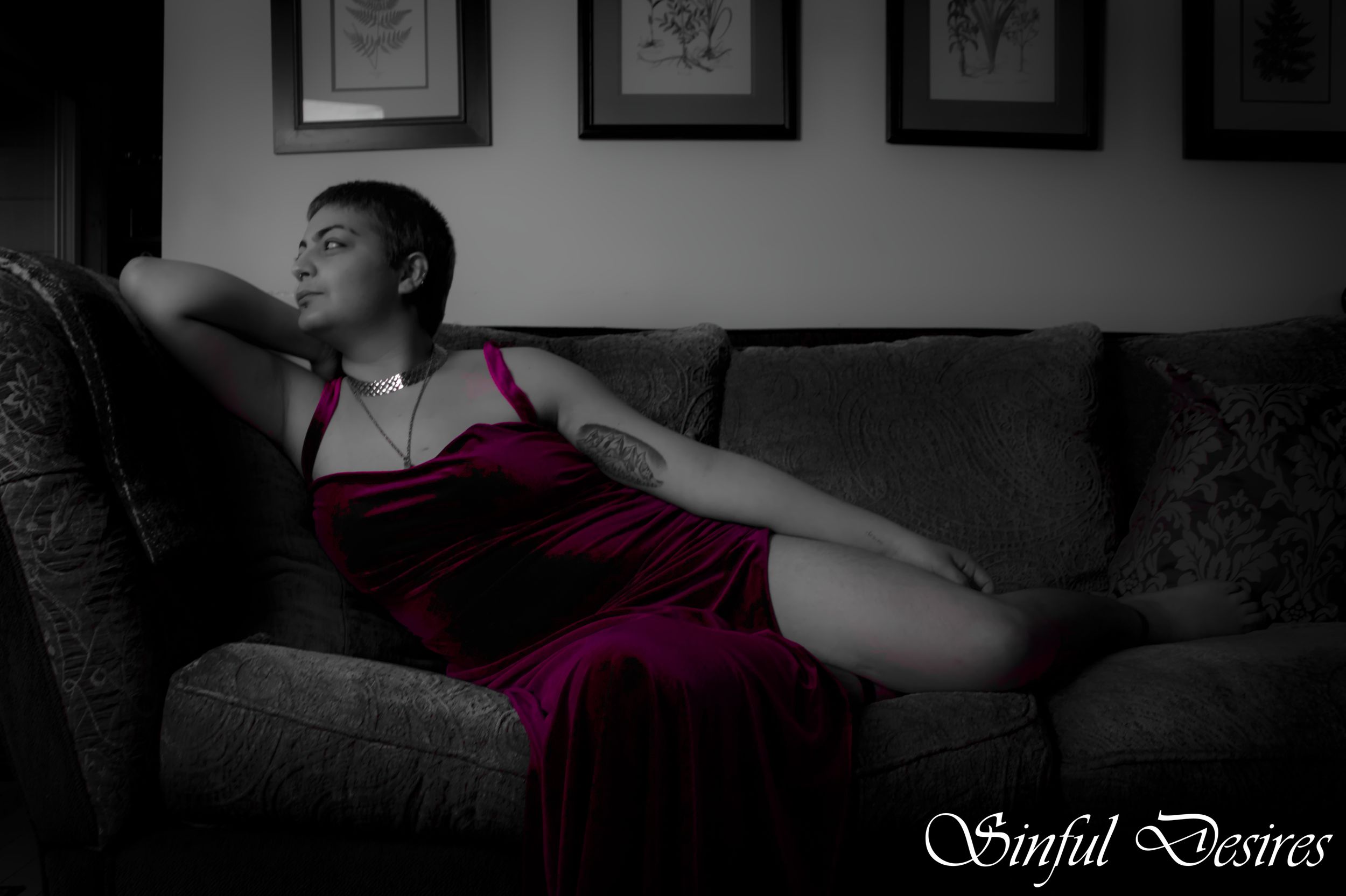 A person in a burgundy dress reclines on a dark couch beneath framed artwork in dramatic black and white lighting.