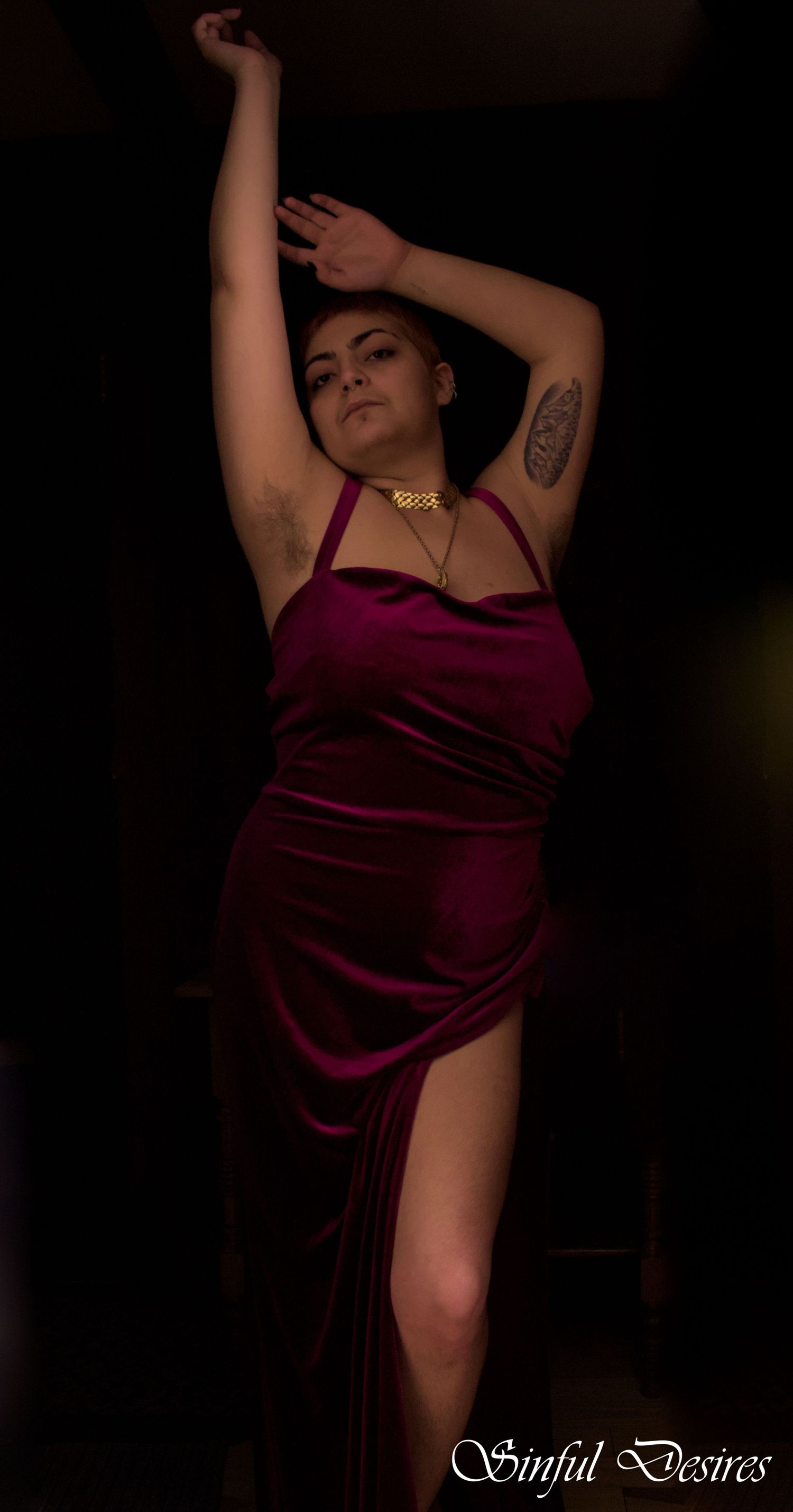 Figure in burgundy velvet dress poses dramatically in low light.