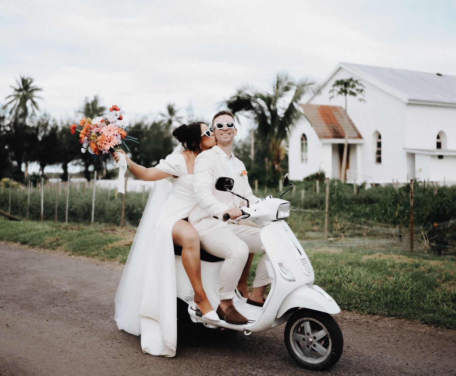 White Vespa scooter parked on country road with wedding couple and white church in background among palm trees.