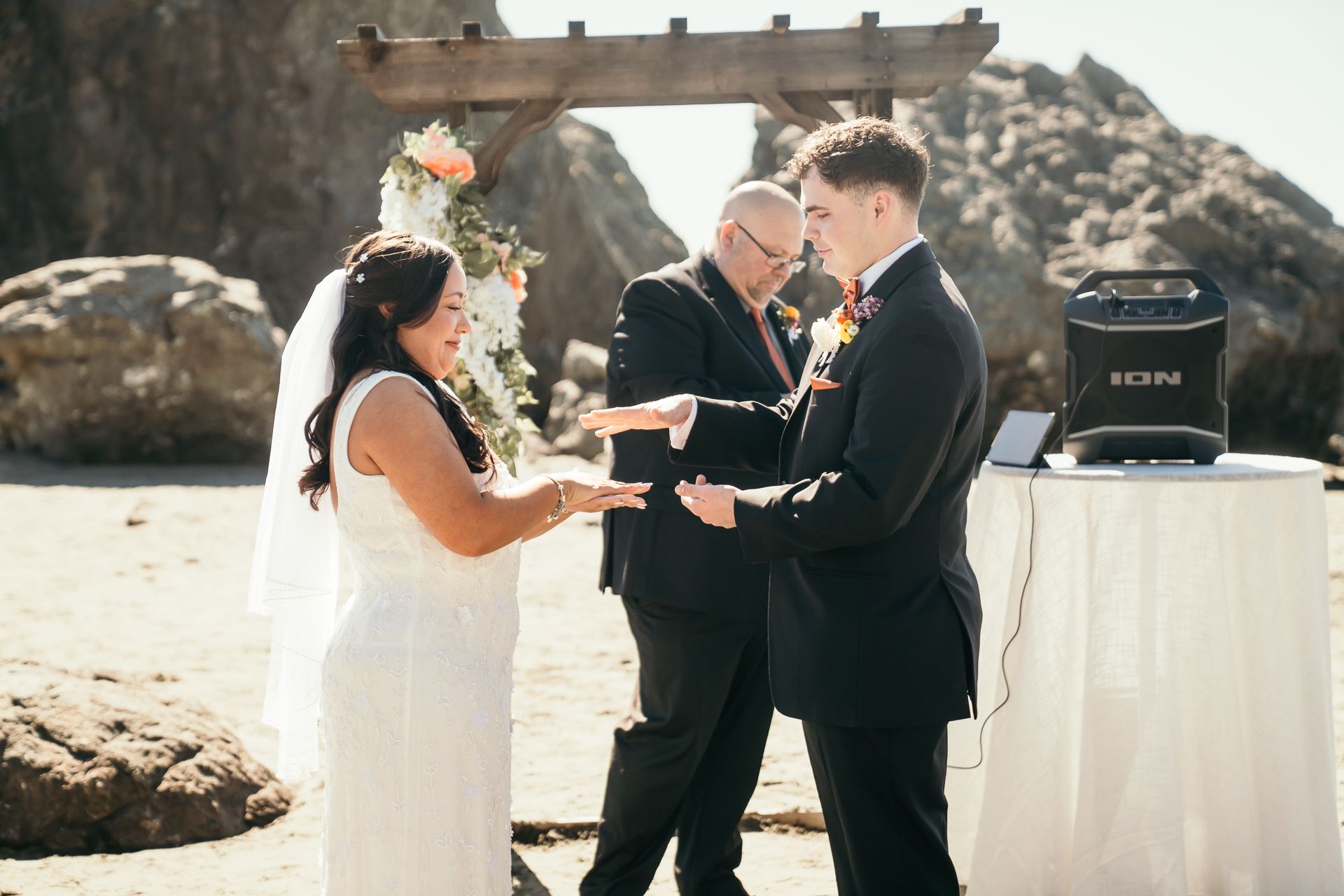 Couple exchanging rings during an outdoor beach wedding ceremony with officiant and floral arch.