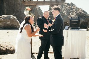 Bride and groom laughing joyfully with officiant during romantic beach wedding ceremony outdoors.