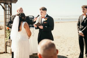 Groom reads vows to bride during outdoor beach wedding ceremony with officiant and groomsmen.
