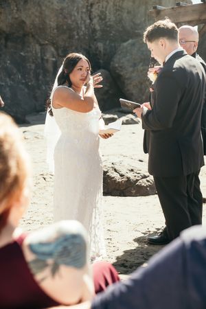 A bride in a white lace gown reads her vows to her groom during a romantic beach wedding ceremony.