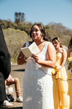 A bride in a white floral gown reads vows from a book during an outdoor wedding ceremony with bridesmaids.