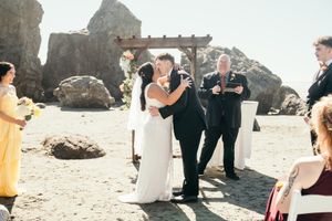 Bride and groom share first kiss at outdoor beach wedding ceremony with rocky coastline backdrop.