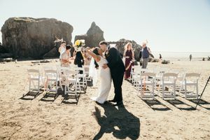 Newlyweds share their first kiss on a sunny beach wedding ceremony with white chairs and rock formations.